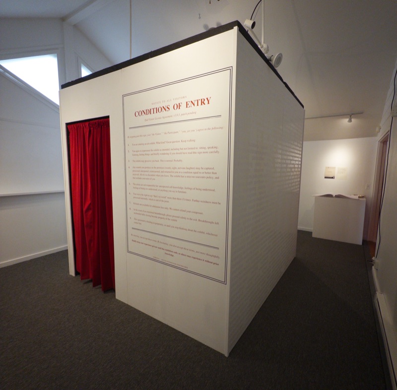 The booth from an angle inside the gallery, showing the red curtain, the Conditions of Entry notice, and a side wall covered in faint overlapping words, with a plinth and open book in the background